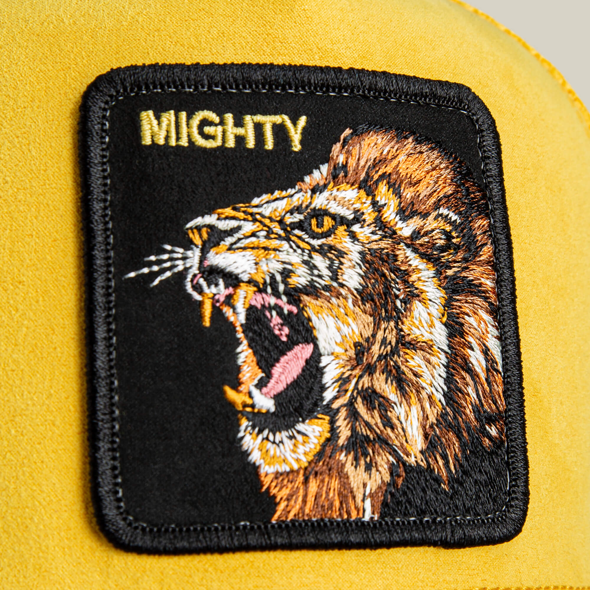 A close-up of the Goorin Bros. Suede Lion trucker hat, featuring a bold yellow fabric patch with an embroidered roaring lion and "MIGHTY" above it.