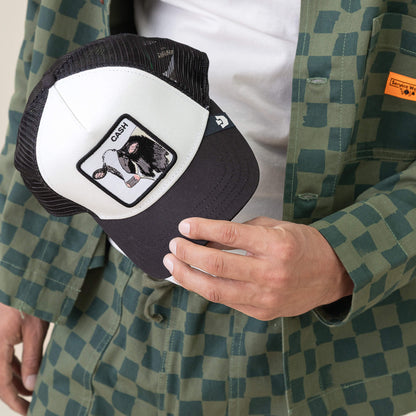 A person in a green checkered jacket holds the Goorin Bros. Cash Cow Trucker hat, featuring a black and white design with a bold bovine cash register patch labeled CASH—capturing the spirit of the Cash Cow.