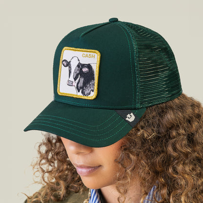 A person wears the Goorin Bros. Cash Cow Trucker, a dark green mesh cap featuring a patch with a bold cow graphic and “Cash Cow” text.
