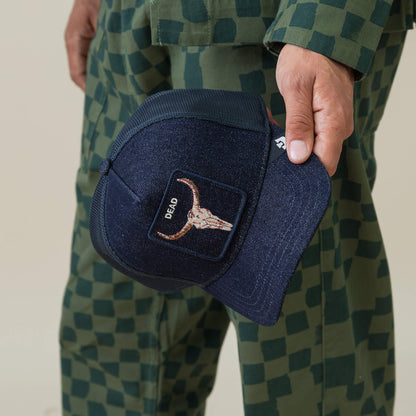 A person in a green checkered outfit holds the Goorin Bros. Denim Dead cap—a navy blue biker hat featuring a bull skull embroidery patch and the word "DEAD.