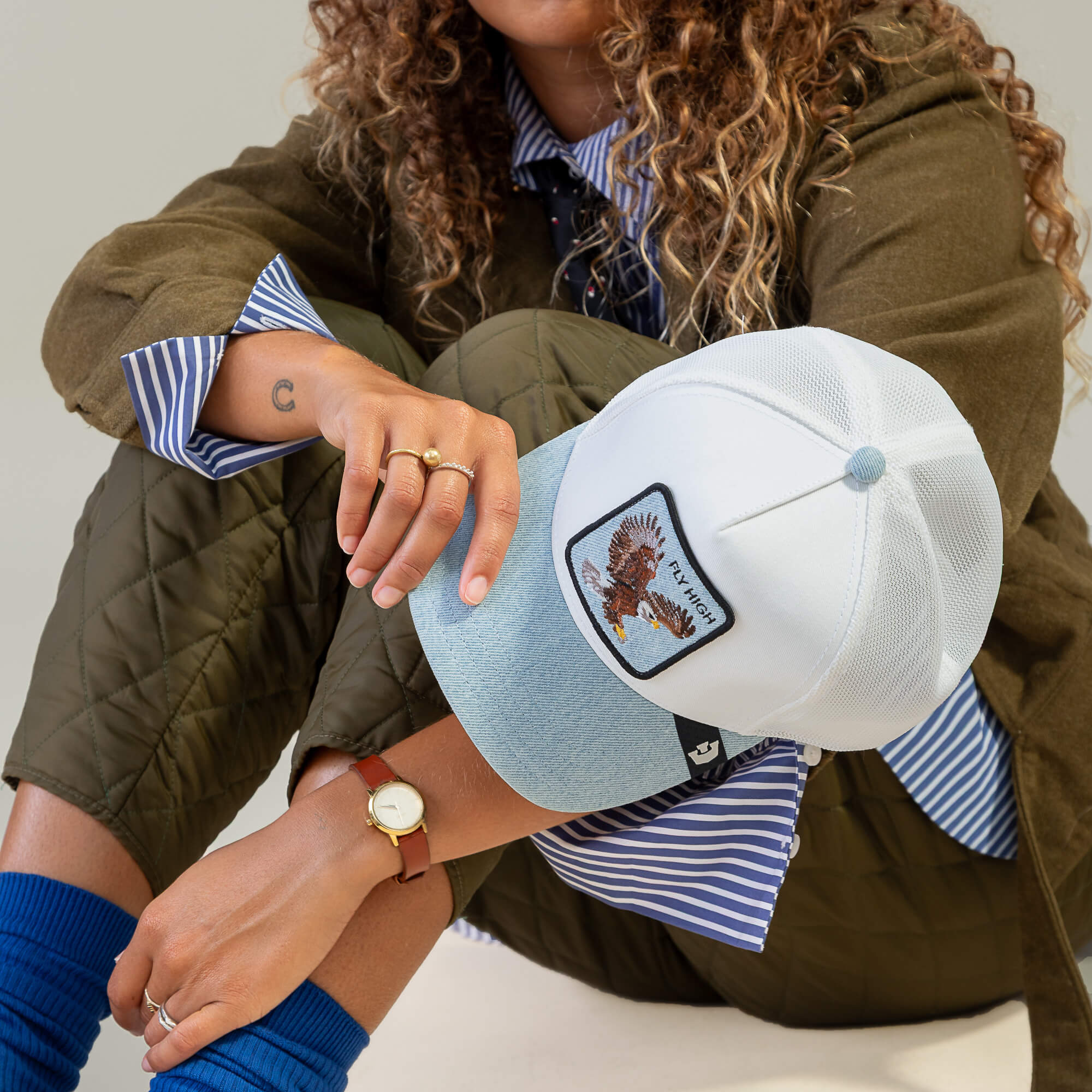 Person seated cross-legged, sporting the Goorin Bros. Denim Fly High cap with eagle patch, olive quilted jacket, blue striped shirt and socks, and a watch. True altitude junkie vibe; face is partially obscured.