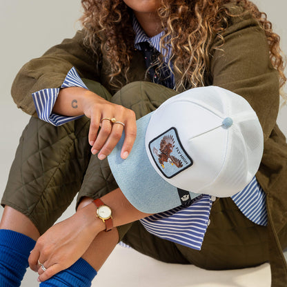 Person seated cross-legged, sporting the Goorin Bros. Denim Fly High cap with eagle patch, olive quilted jacket, blue striped shirt and socks, and a watch. True altitude junkie vibe; face is partially obscured.