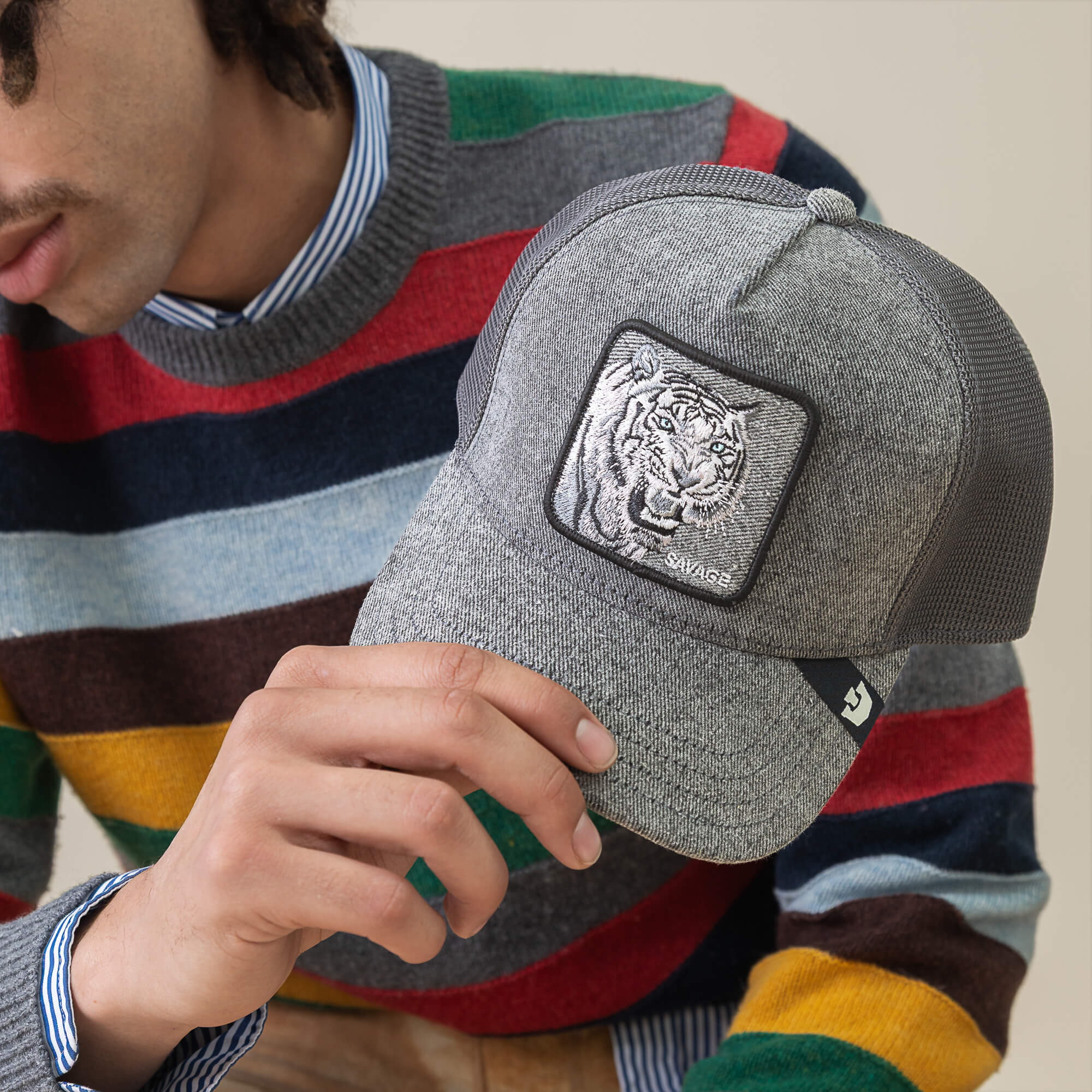 A person in a multicolored striped sweater holds the Goorin Bros. Denim Savage cap, a gray streetwear hat featuring feral embroidery with an embroidered tiger patch on the front.