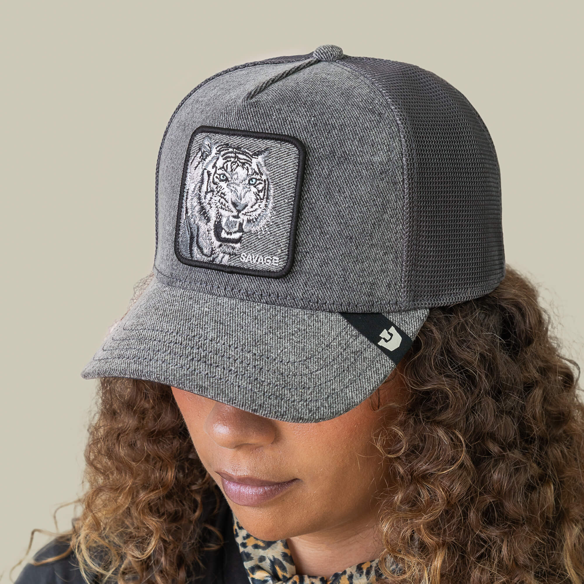 A person with curly hair wears the Goorin Bros. Denim Savage cap, a gray streetwear piece with a tiger patch and "SAVAGE" embroidered on the front.