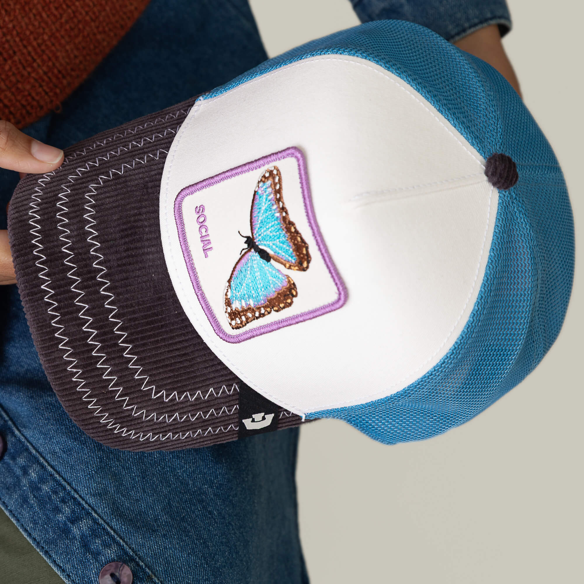 A person holds the Goorin Bros. Social Remix hat, featuring a blue butterfly patch and the word SOCIAL on the front panel, with blue mesh sides and a dark corduroy brim.