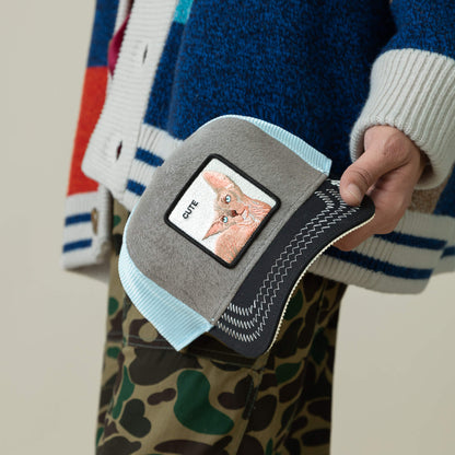 A person in camouflage pants holds the Goorin Bros. Cute Remix—a gray and blue cap with a patch of a hairless cat’s face and "CUTE" above it.