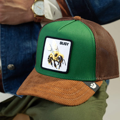 A vibrant green and honey brown Goorin Bros. Busy Remix hat features a mesh design, bee patch, and BUSY on the front. Styled here with a denim jacket and watch, it’s the perfect statement cap for any garden-party look.