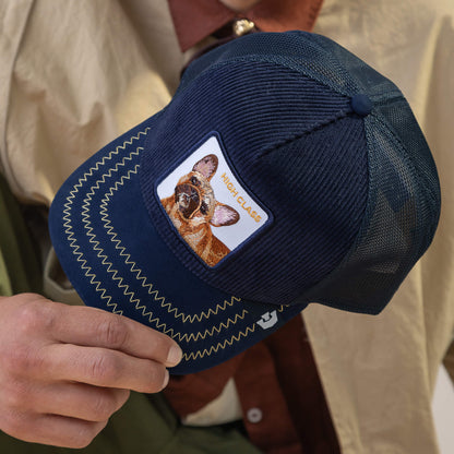A person holds the Goorin Bros. High Class Remix cap, a navy blue mesh hat with an embroidered dog patch and "HIGH CLASS" text, its navy textures adding depth.