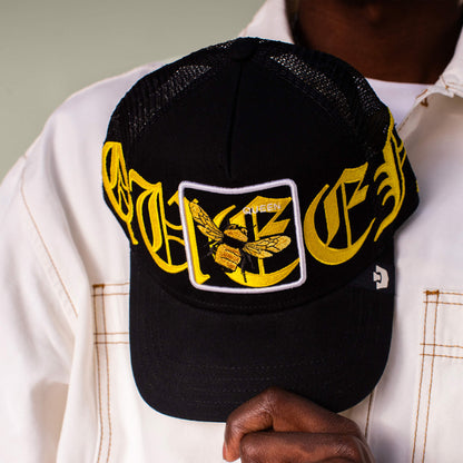 A person holds the Goorin Bros. Old English Queen black mesh cap, featuring striking black and yellow hues, Old English lettering, and a patch with a yellow moth and QUEEN text.