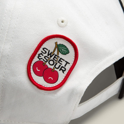 A close-up of the Goorin Bros. Pop Appeal white cap featuring a cherry patch with two cherries, a green leaf, and red-outlined SWEET & SOUR text.