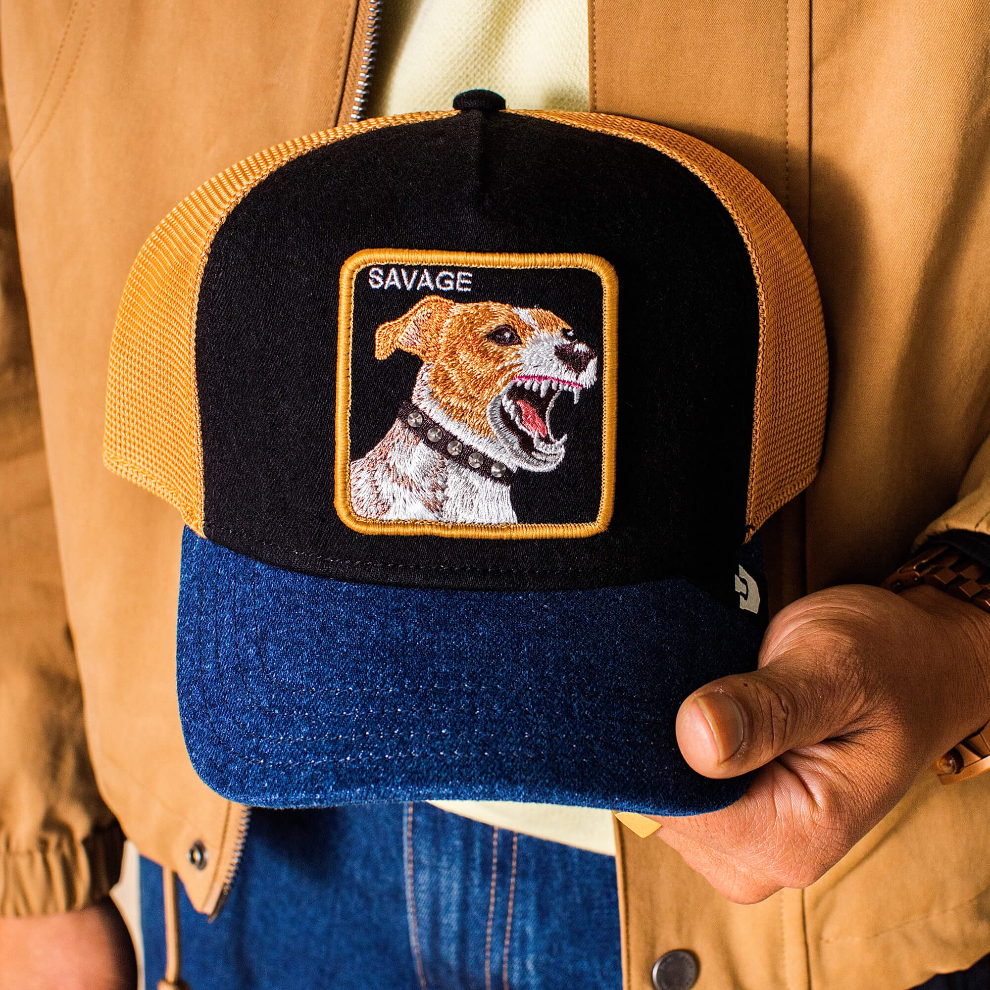 A person holds the Goorin Bros. Savage Dawg cap, showcasing an embroidered snarling dog and "SAVAGE" text on a bold mixed denim design.