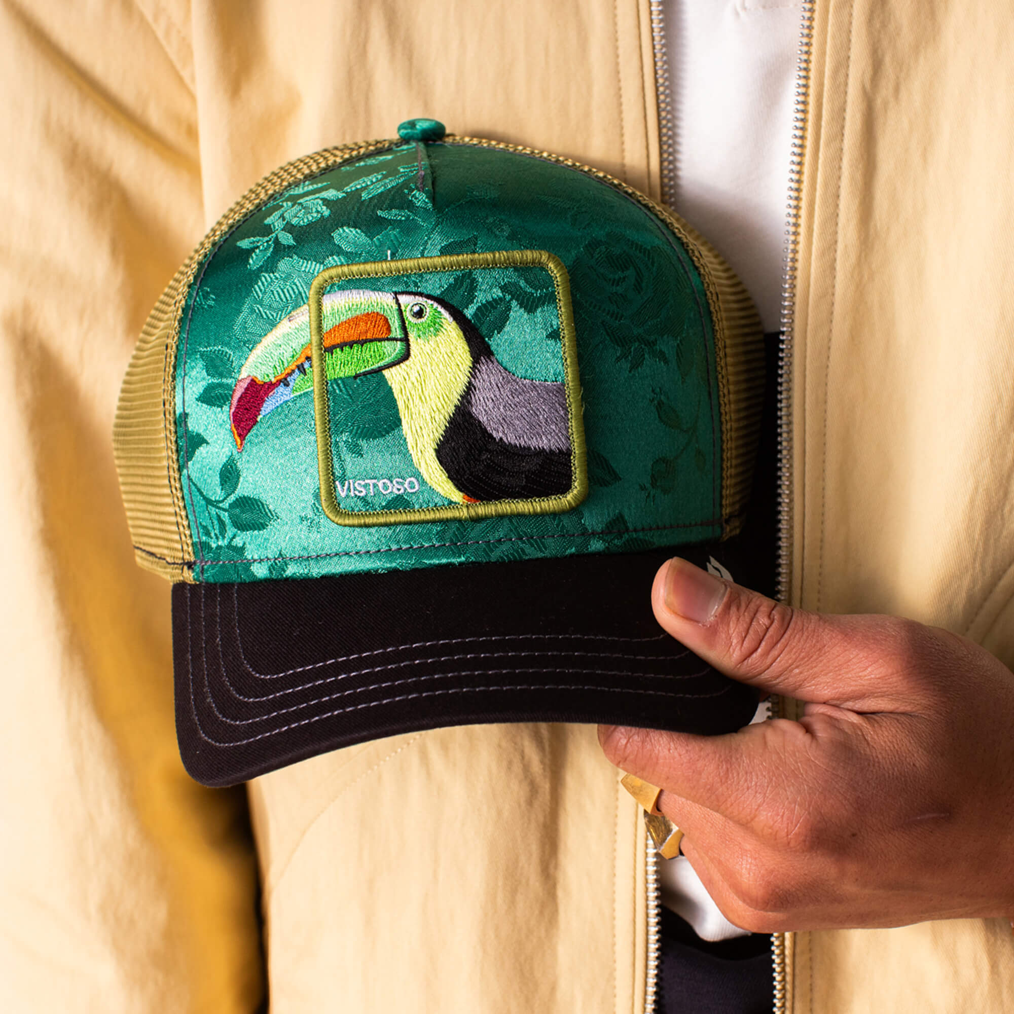 A person holds the Goorin Bros. Belize Toucan trucker hat, a green cap with an embroidered toucan patch and VISTO60 on the front, inspired by the vibrant Belize Toucan.