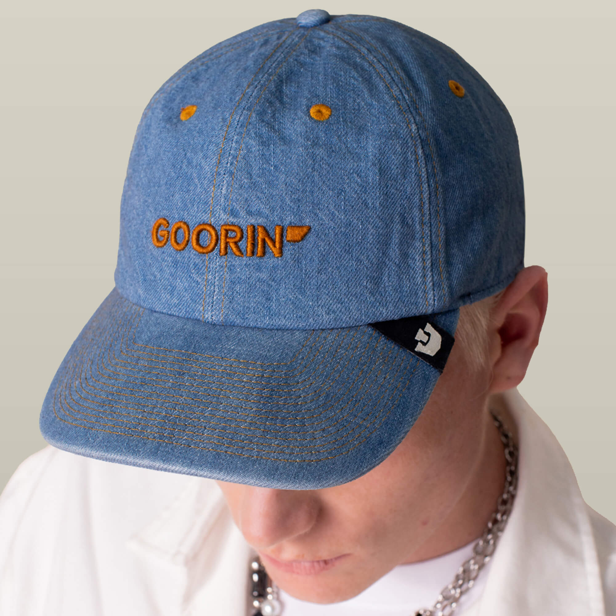 A person wearing the Goorin Bros. Papa Core Denim hat—a low-profile blue denim cap with orange "GOORIN" embroidery—pairing it with a white shirt and a chunky chain necklace.