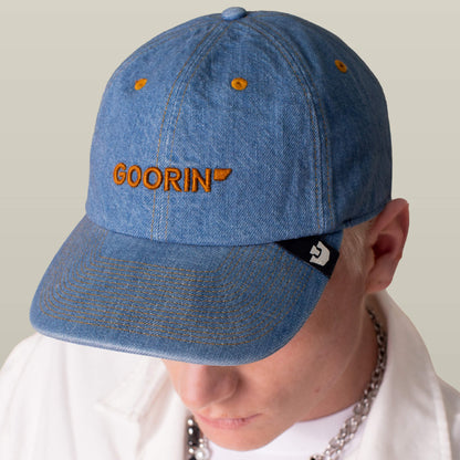 A person wearing the Goorin Bros. Papa Core Denim hat—a low-profile blue denim cap with orange "GOORIN" embroidery—pairing it with a white shirt and a chunky chain necklace.