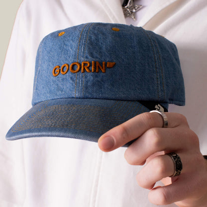 A person holds the Papa Core Denim hat by Goorin Bros., featuring a blue denim design and orange GOORIN embroidery on the front, while wearing a white top and silver rings.