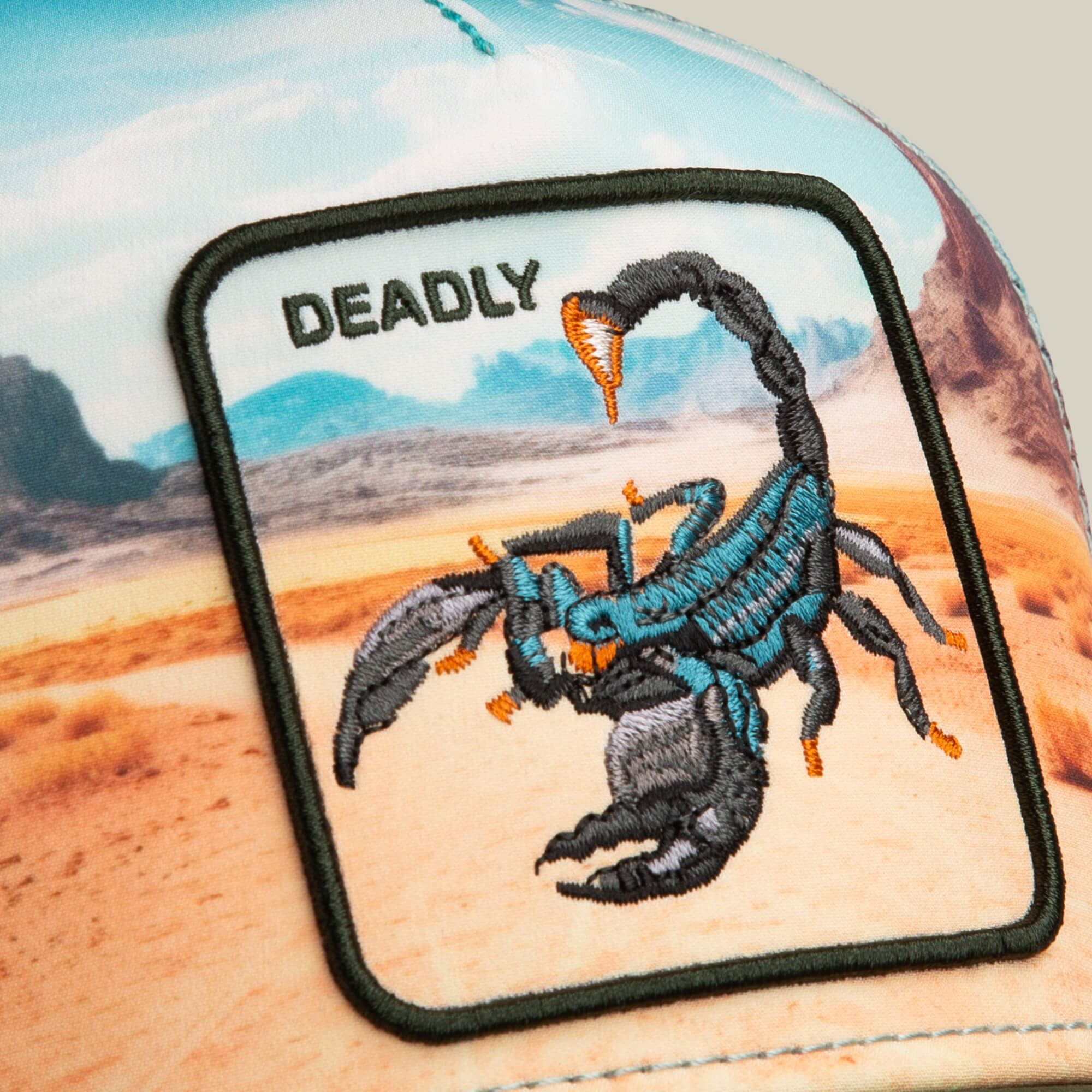 A close-up of the Goorin Bros. "Scorpion In The Element" trucker hat, featuring an embroidered scorpion patch with "DEADLY" above it, set against a desert landscape background.