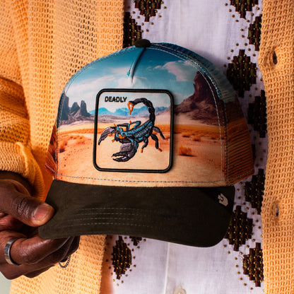 A person holds the Goorin Bros. Scorpion In The Element trucker hat, featuring a DEADLY patch and desert landscape design on the front with a breathable mesh back.