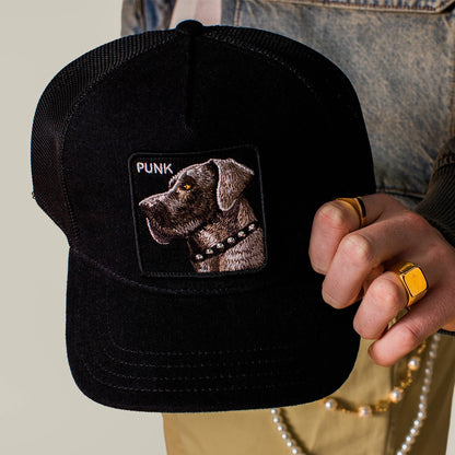 A person holds the Goorin Bros. Punk Dawg black trucker hat, which features a patch with an embroidered dog and "PUNK." Gold rings and a classic denim jacket complete the edgy style.