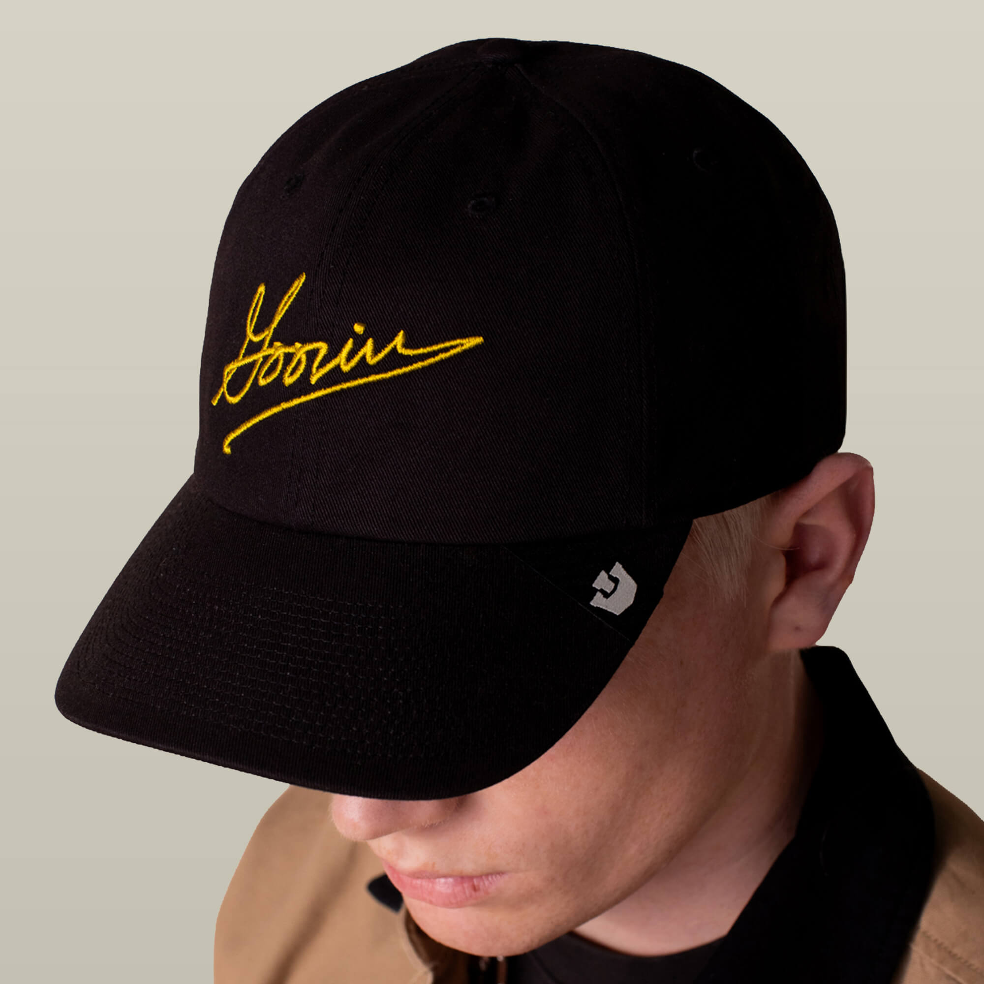 A person wearing a black Goorin Script Baseball cap by Goorin Bros., featuring yellow embroidered branding and a small white logo on the brim, facing slightly downward.