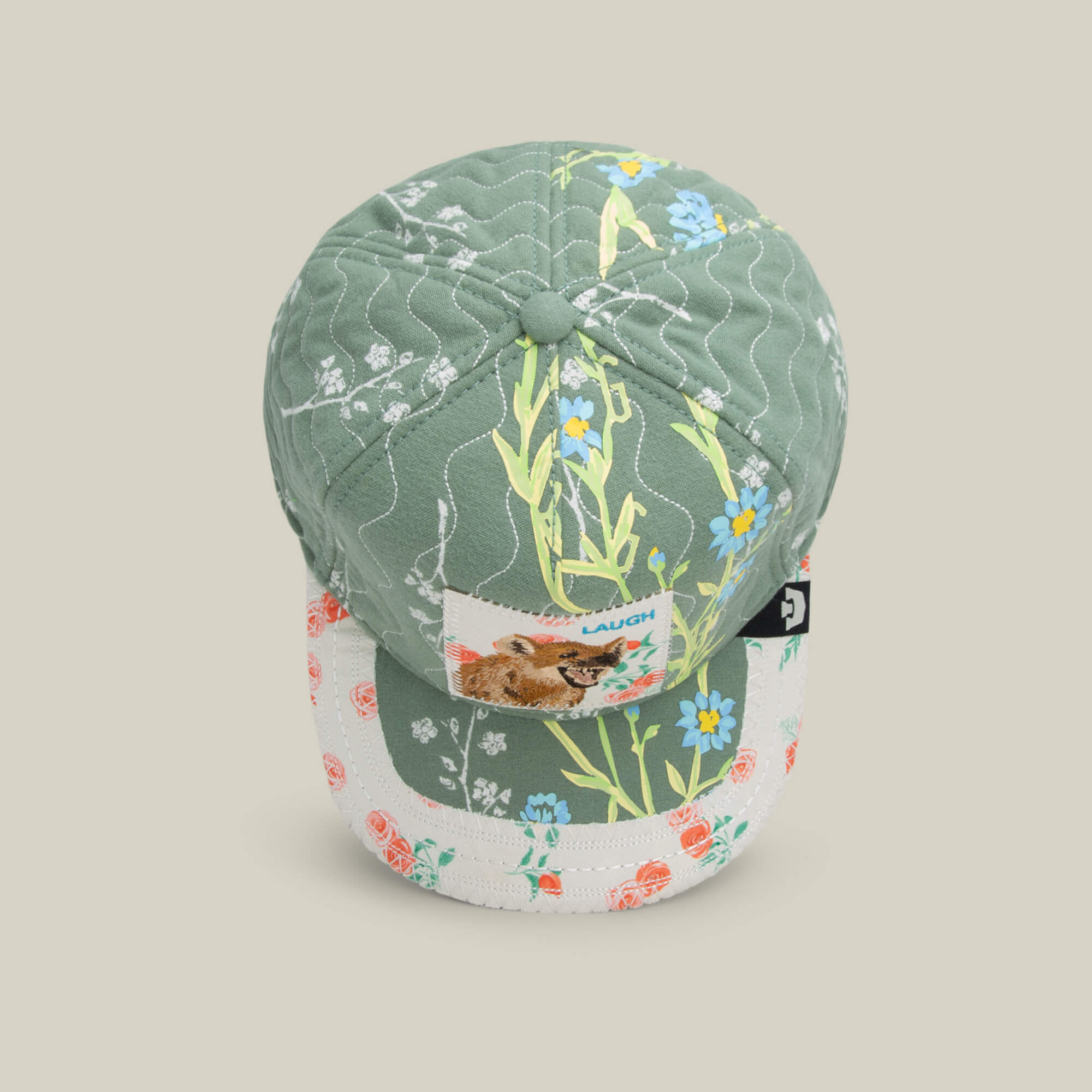 The Goorin Bros. Vol. 1 - Quiltbag: Laugh is a green and white floral trucker hat with patchwork patterns, featuring a front patch of a dog and the word “LAUGH.”.