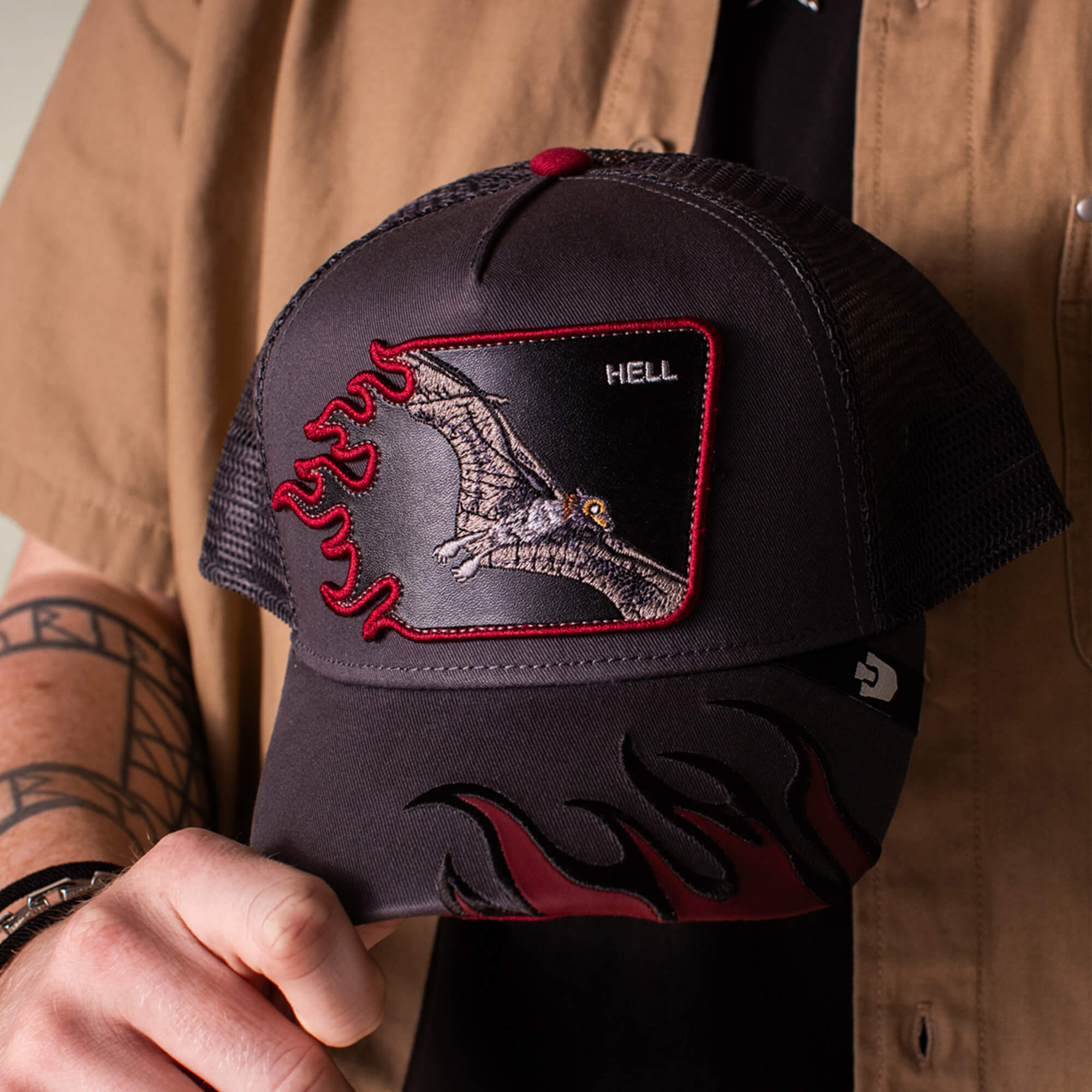 A person holds the Goorin Bros. Hell Flames charcoal trucker hat, featuring a pterodactyl patch labeled HELL, bold red flame designs, and mesh back panels.