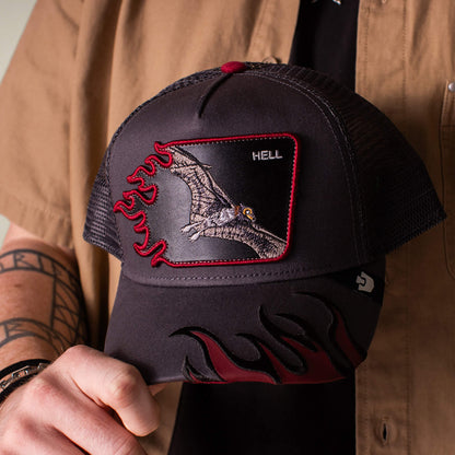 A person holds the Goorin Bros. Hell Flames charcoal trucker hat, featuring a pterodactyl patch labeled HELL, bold red flame designs, and mesh back panels.