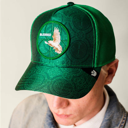 A person wears the Religulous - Blessed cap by Goorin Bros., featuring a deep green color, flying bird patch, “BLESSED” on the front, textured pattern, and a logo tag on the side for stylish detail.