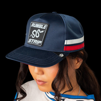 A person in a Goorin Bros. Pole Position - Rumble Strip cap, featuring red, white, and blue stripes and a signature patch, looks downward against a black background—a style popular among drift drivers.
