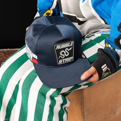 A person in green and white striped pants and a colorful top holds the Goorin Bros. Pole Position - Rumble Strip cap—navy blue with a Rumble Strip patch, ideal for drift drivers seeking standout style.