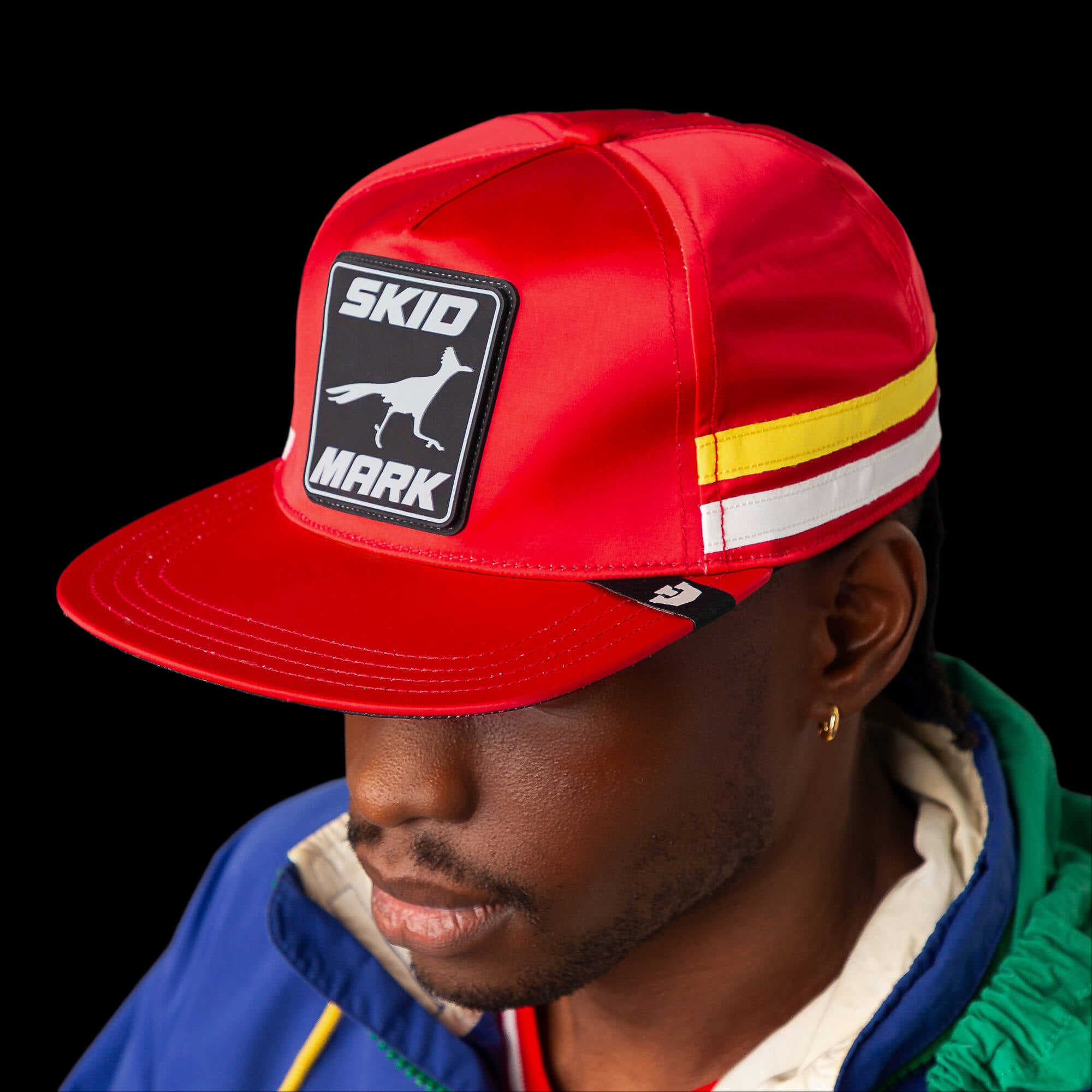 A person in a red Goorin Bros. "Pole Position - Skid Mark" cap, featuring a bird graphic, and a blue-green jacket stands against a black background, embodying the spirit of roadrunner speed.