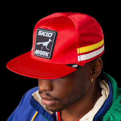 A person in a red Goorin Bros. "Pole Position - Skid Mark" cap, featuring a bird graphic, and a blue-green jacket stands against a black background, embodying the spirit of roadrunner speed.