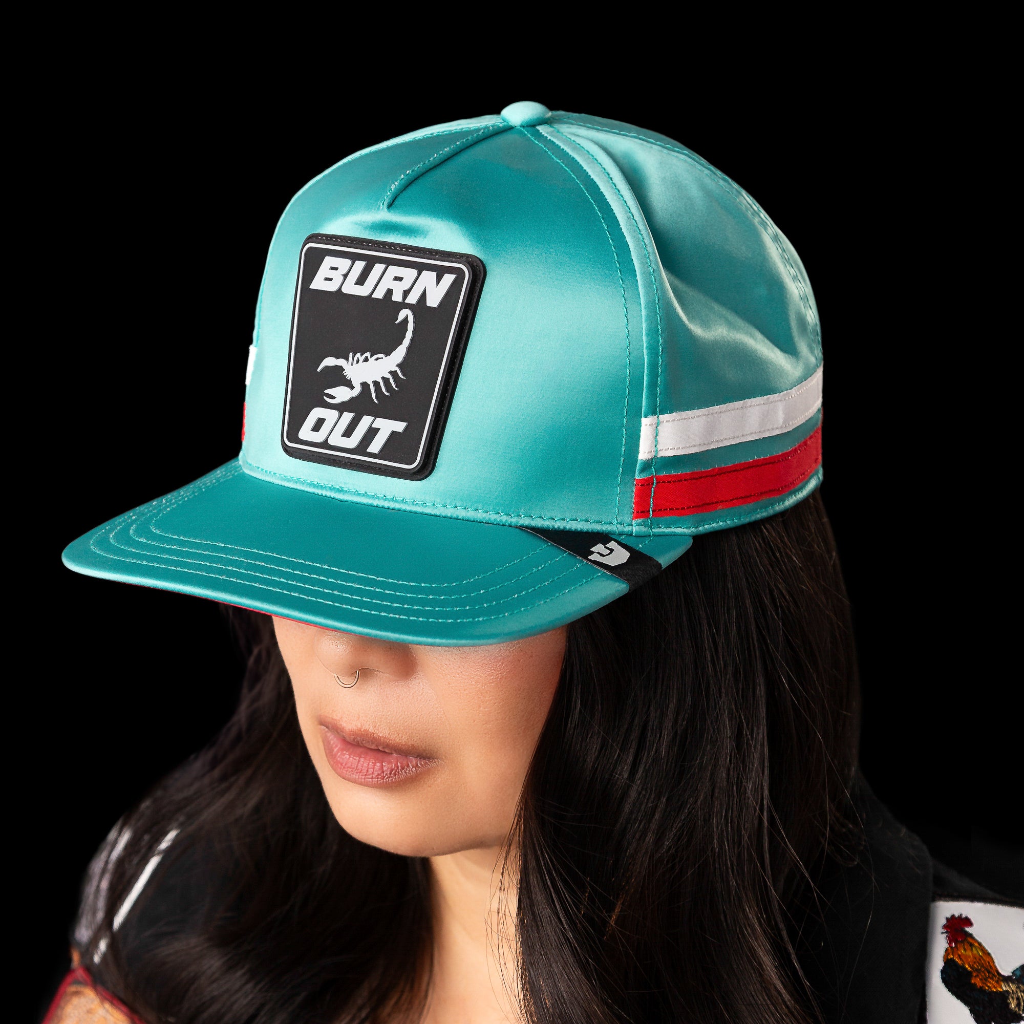 A person wears the Goorin Bros. Pole Position - Road Kill teal racing cap featuring a BURN OUT patch, scorpion graphic, red and white side stripes, and a nose ring, set against a black background.