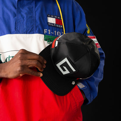 A person in a Tommy Hilfiger jacket holds a black Goorin Bros. Pole Position - Road Kill hat featuring a stylized G logo on the front, all set against a sleek black background.