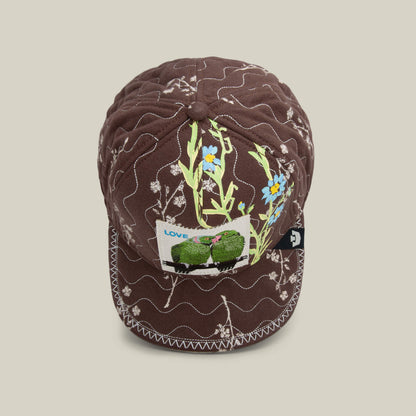 The Goorin Bros. Vol. 1 - Quiltbag: Love is a hand-stitched OG Trucker cap in chocolate brown quilting with white stitching, yellow and blue floral embroidery, and a “LOVE” patch featuring two green frogs.
