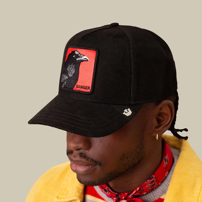 A sharp-eyed individual wears a Goorin Bros. black cap featuring the Corvo Rosso signal red patch with a crow and "DANGER," paired with a yellow jacket, red patterned scarf, and gold hoop earring on a beige background.