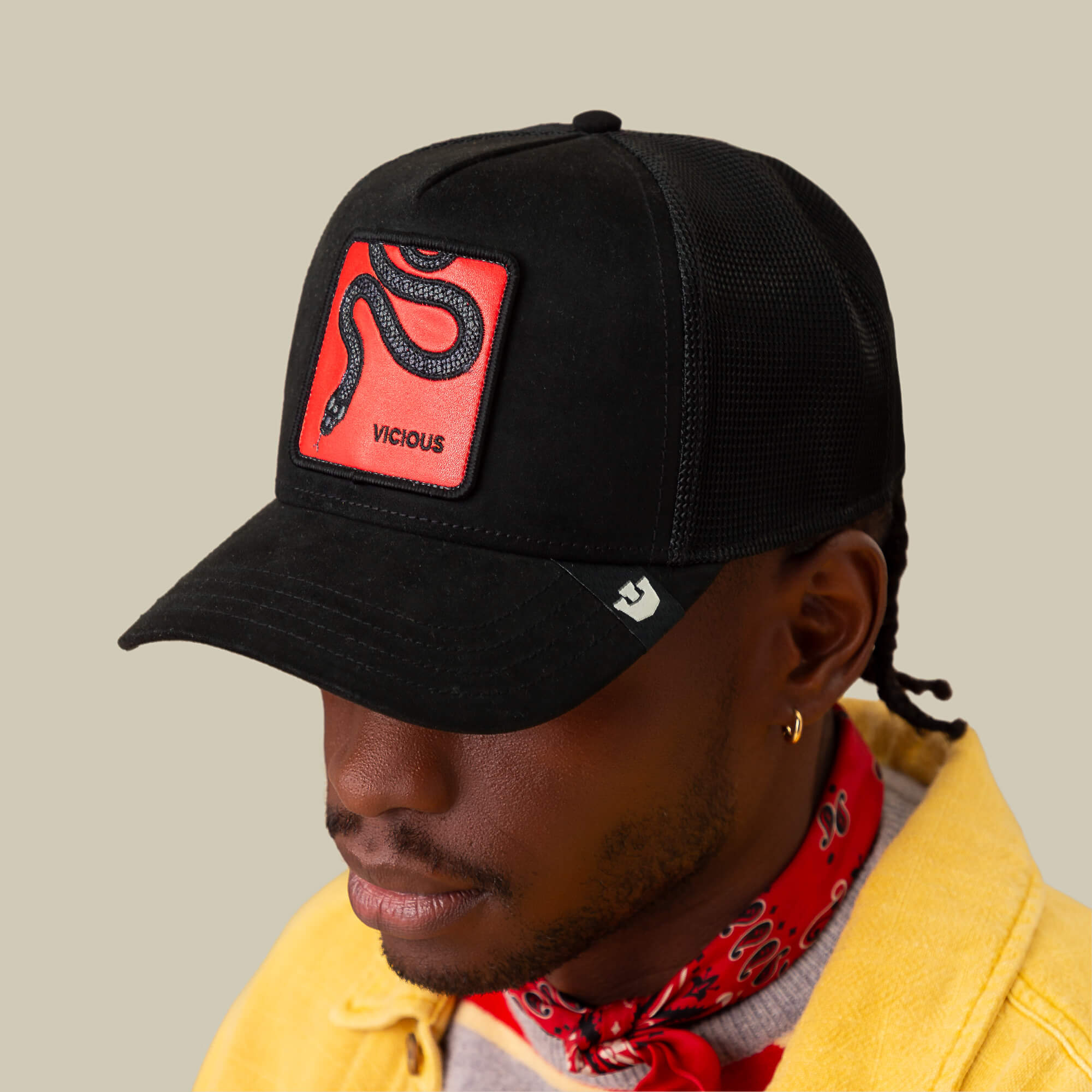 A person wears a Goorin Bros. Serpente Rosso black microsuede cap with VICIOUS patch, paired with a yellow jacket and red bandana, standing against a beige background.