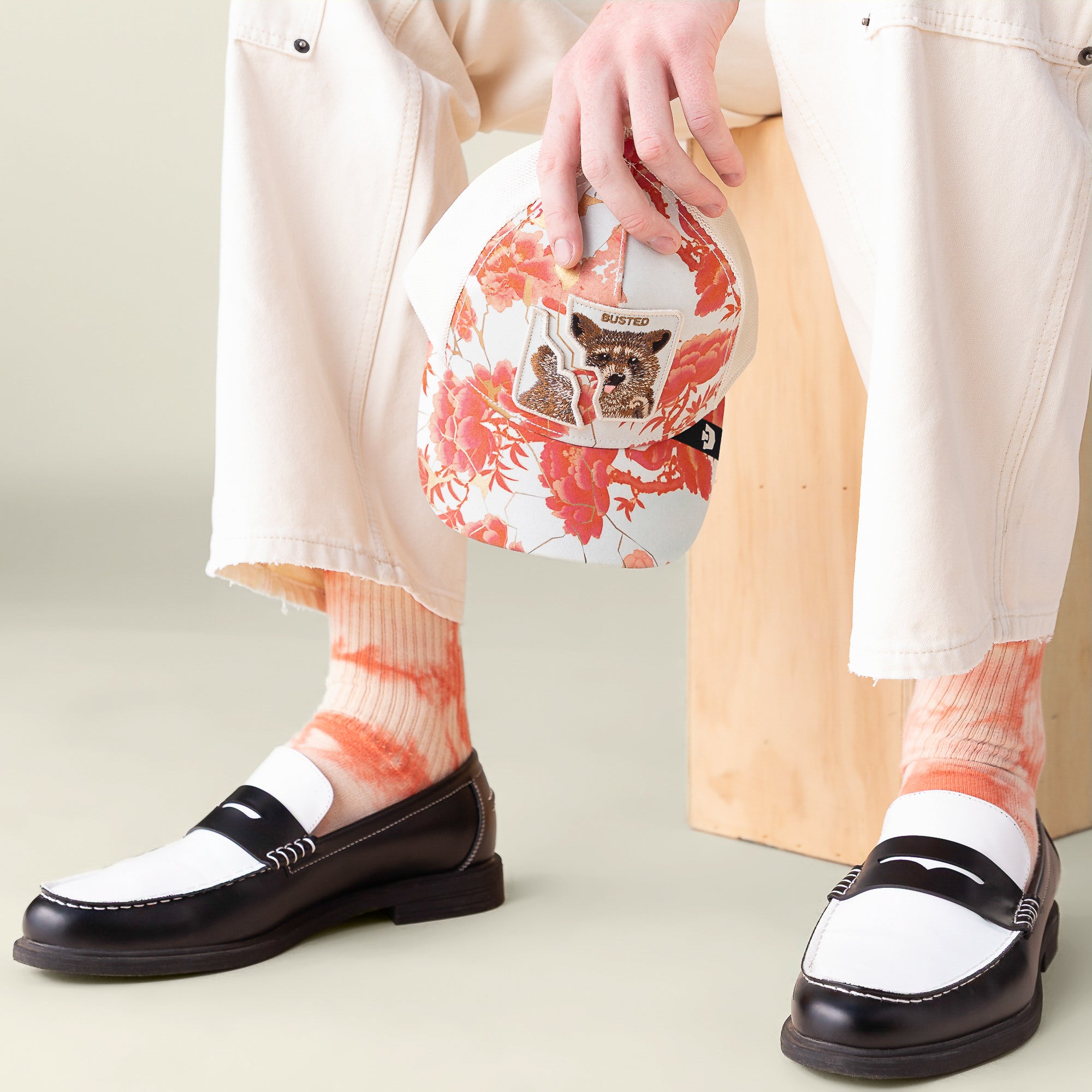 A person sits on a light wooden block, holding the Goorin Bros. “Break The Law” cap featuring floral and animal patches, paired with cream pants, orange tie-dye socks, and black-and-white loafers.