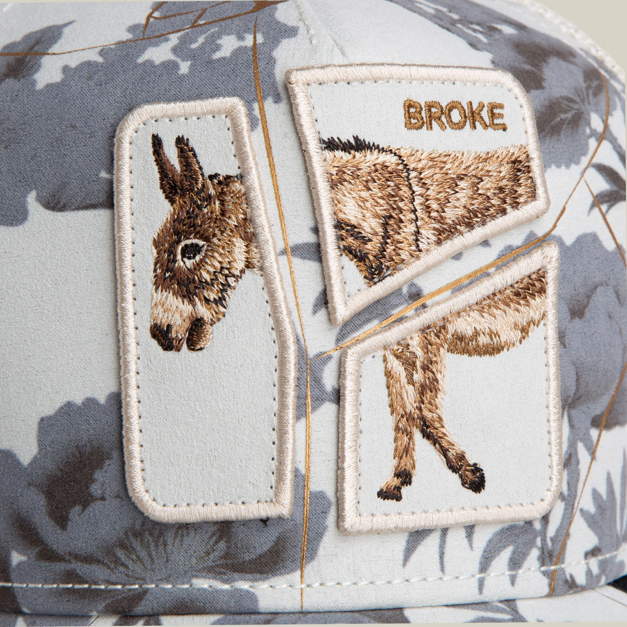 The Goorin Bros. "Break The Bank" cap features embroidered patches with a broken donkey image and "BROKE" above it, set against a gray and white abstract background—an ideal novelty gift for anyone short on cash.