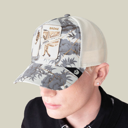 A person with pale skin and platinum blond hair wears a mesh-back Goorin Bros. cap called "Break The Bank," featuring a floral pattern and a horse patch reading “BROKE.” The cap’s brim obscures most of their face.