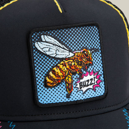 Close-up of Goorin Bros. Oh Bee Hive black trucker hat with an embroidered bee patch and "Buzz!" comic-style speech bubble on a dotted blue pop-art background.