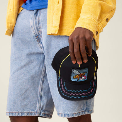 A person in a yellow jacket and denim shorts holds the Goorin Bros. Oh Bee Hive trucker hat, featuring colorful stitching and a playful pop-art taco patch.