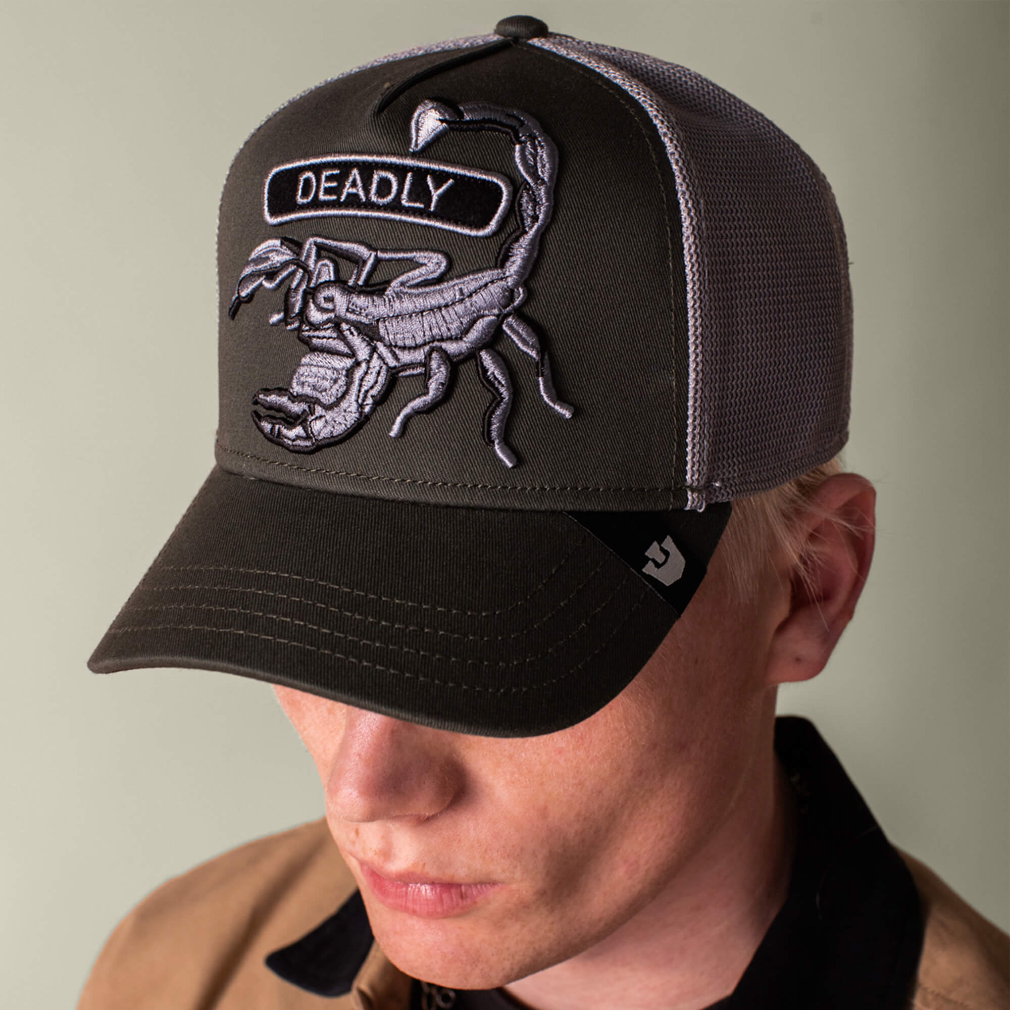 A person wearing the Goorin Bros. Classic Rocker - Deadly, a dark baseball cap with a grey mesh back and a scorpion patch labeled “DEADLY” on the front.