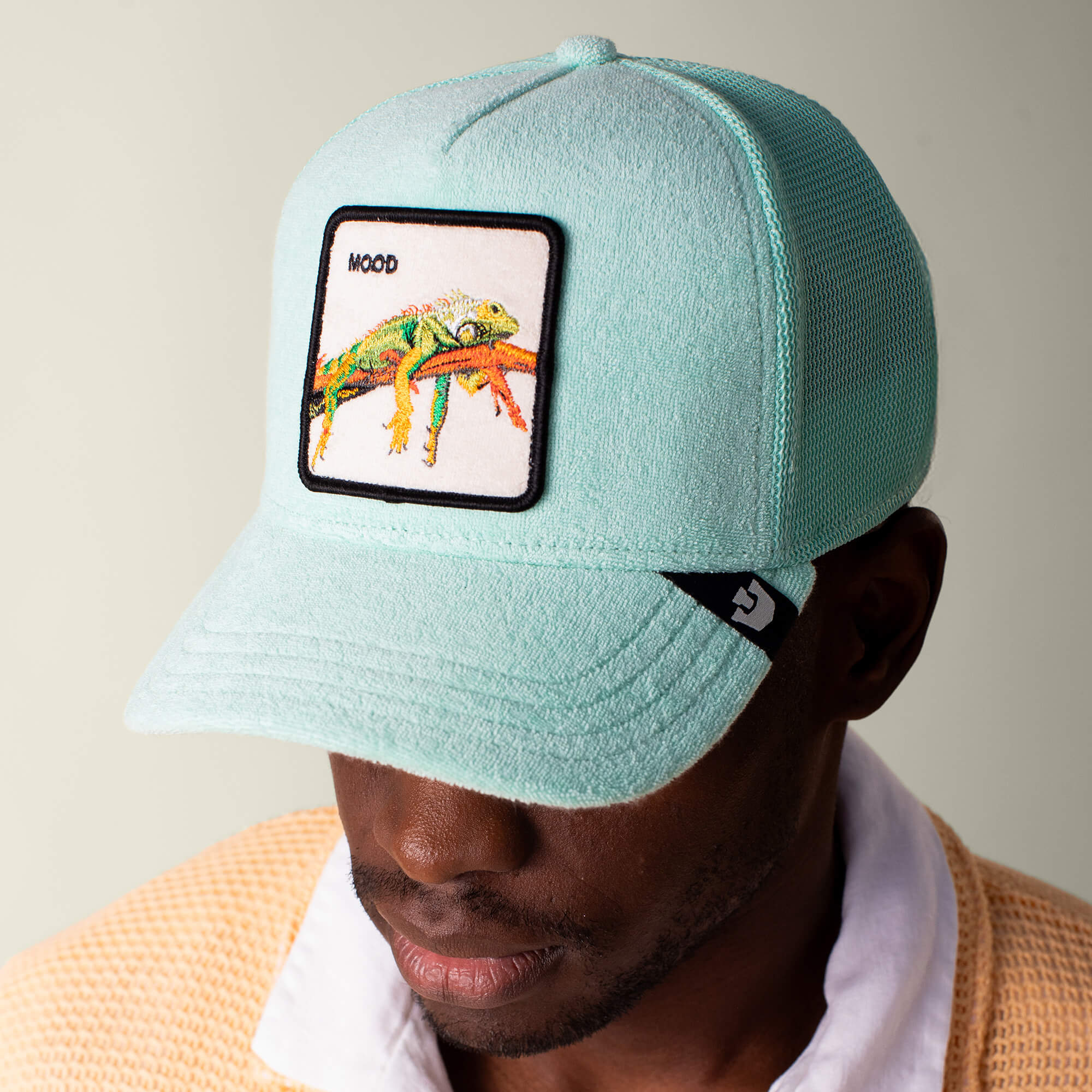 A person wearing Goorin Bros. "Terry Cloth Iguana"—a light blue, comfortable cap with a lizard patch labeled MOOD—looks downward, dressed in a beige textured jacket over a white collared shirt.