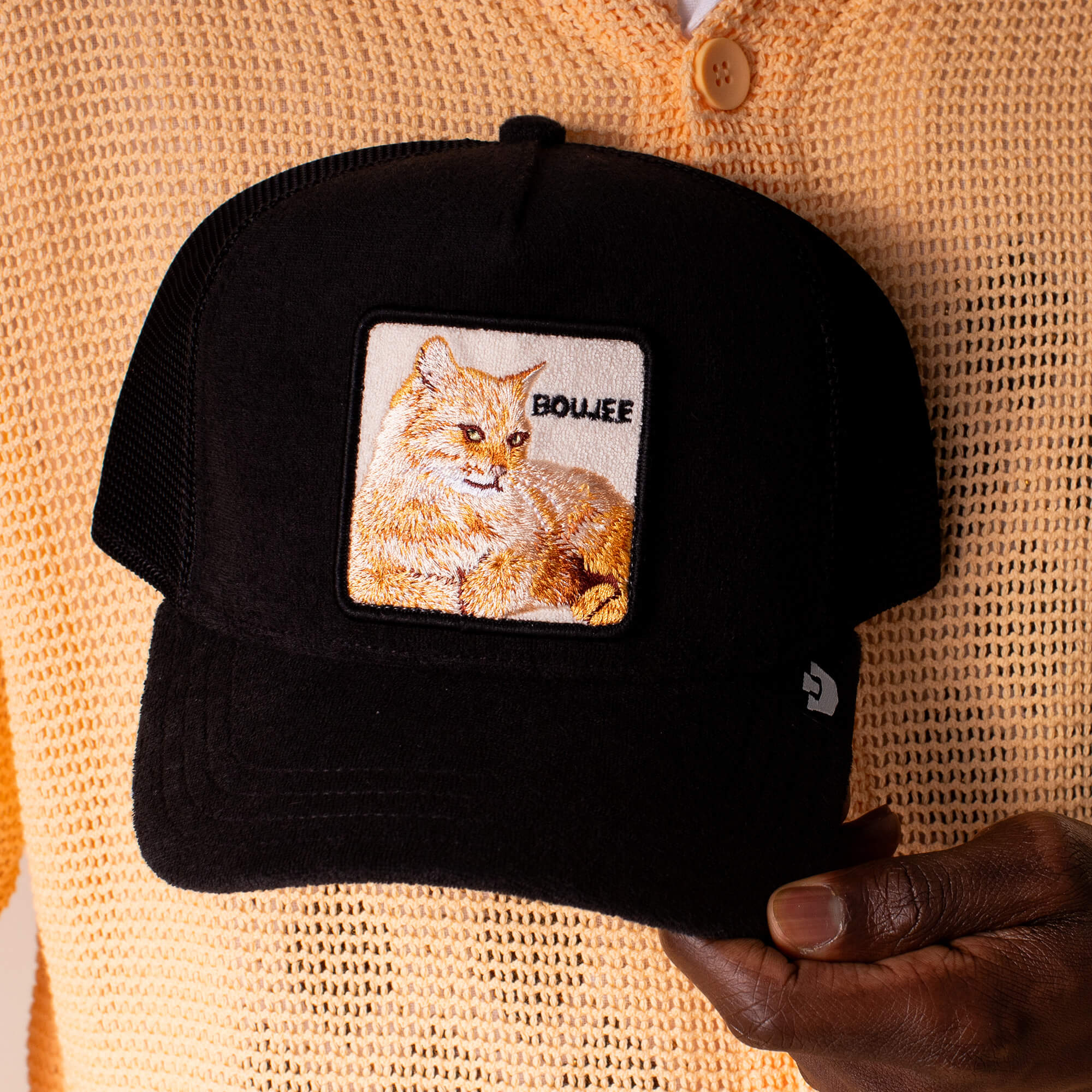 A person in a beige sweater holds Goorin Bros.' Terry Cloth Cat—a plush black terry cloth cap featuring an embroidered cat patch and the word BOUJEE, exuding effortless luxury and lounge-level softness.