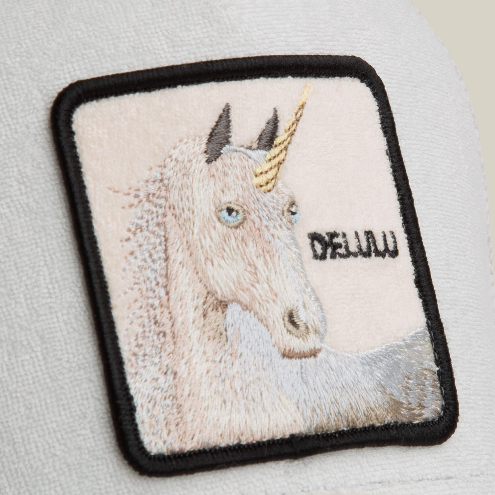 The Terry Cloth Unicorn by Goorin Bros. features a square unicorn patch with a golden horn and blue eyes, labeled DEJA VU, sewn onto soft white terry cloth for a whimsical accent on a light background.