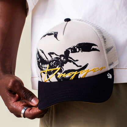 A person holds the Goorin Bros. Outside the Lines Scorpion, a cream and black mesh trucker hat featuring a large black scorpion graphic and "Dagger" embroidered in yellow on the front.