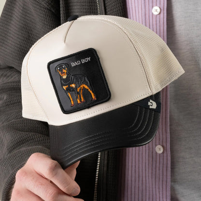 A person holds the Goorin Bros. Faux Leather Rottweiler hat, a beige and black trucker featuring an embroidered patch with a detailed Rottweiler and the words BAD BOY, adding a bold touch to this stylish faux leather accessory.