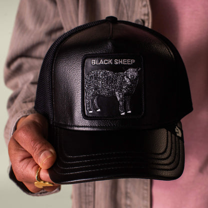 A person holds the Goorin Bros. Faux Leather Sheep trucker hat in black, featuring a faux leather patch with a black sheep and “BLACK SHEEP” text on the front.