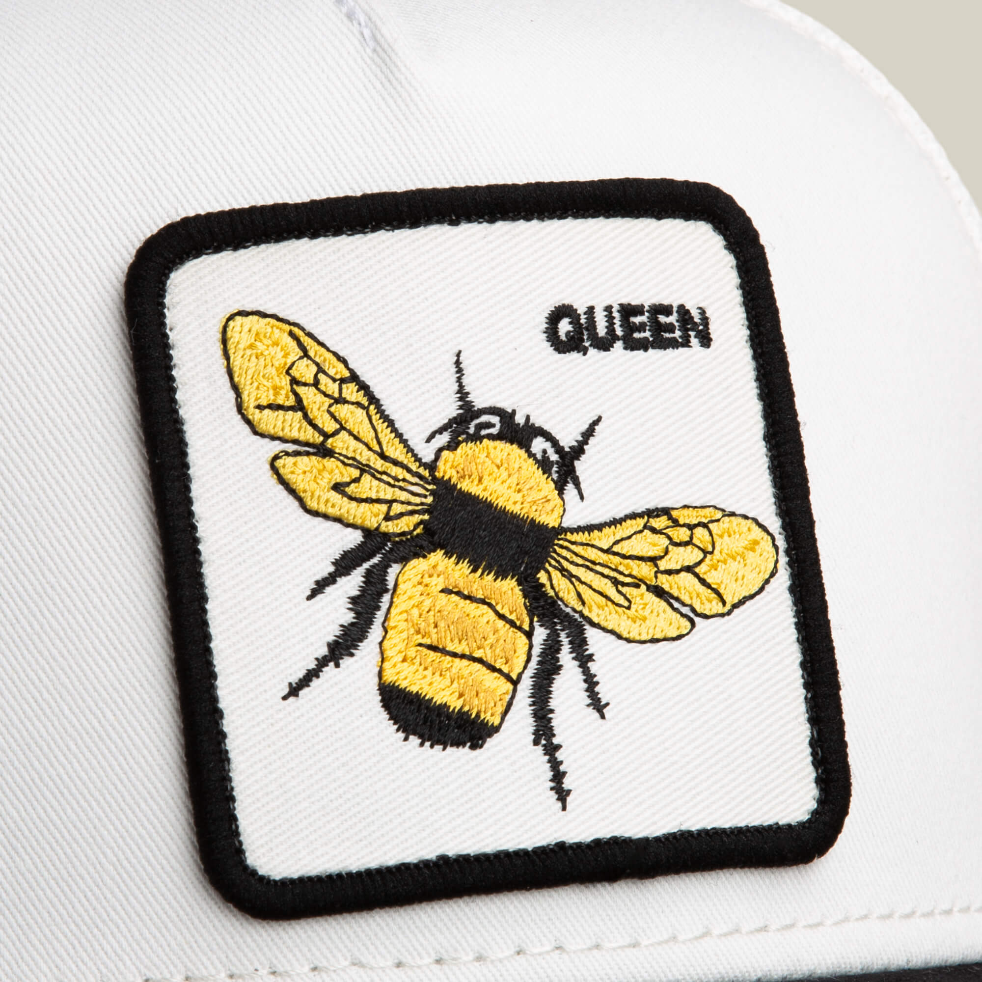 The Goorin Bros. Suede Bee is a white hat with a rectangular Queen patch featuring a yellow and black bee, “QUEEN” text in the corner, and a stylish black microsuede brim.