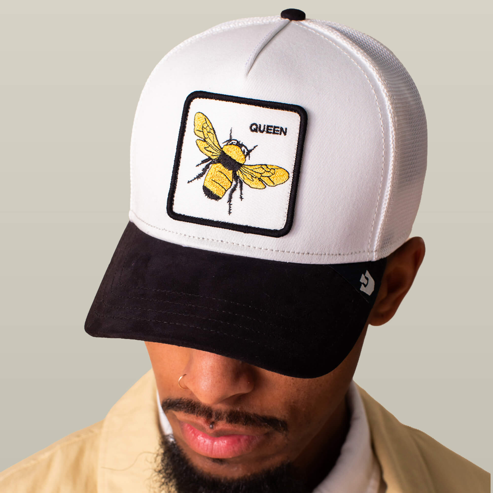 A person wearing the Suede Bee cap by Goorin Bros., featuring a white and black design, a Queen patch with a yellow bee, and a sleek black microsuede brim.